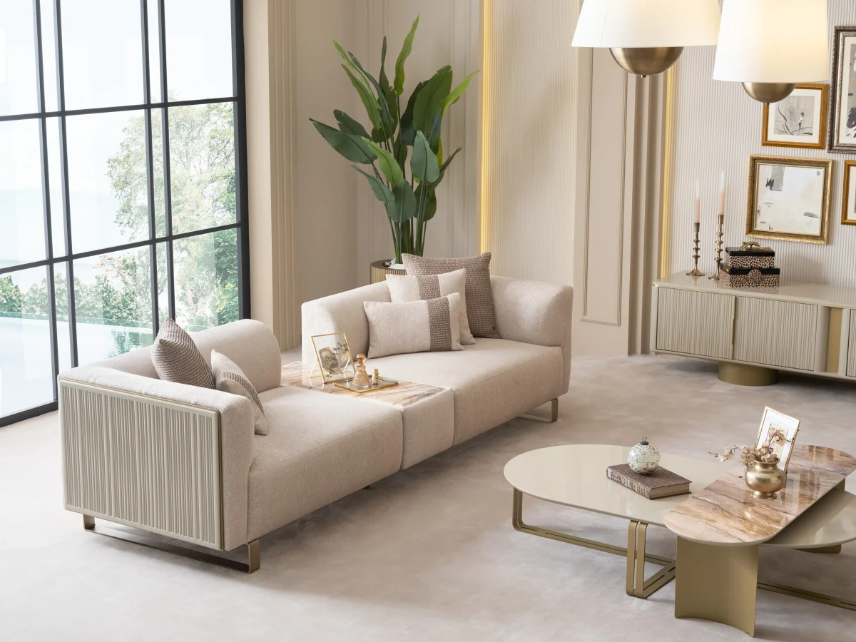 A luxurious modular Grandeur sofa set in a bright, modern living room. The set features cream-colored fabric upholstery with rectangular cushions and sleek gold metallic legs. Beside the modular sections sits a matching rounded armchair with gold accents. The scene includes minimalist decor, a marble coffee table, and large windows, highlighting the elegant design of the Grandeur furniture from Turkey.