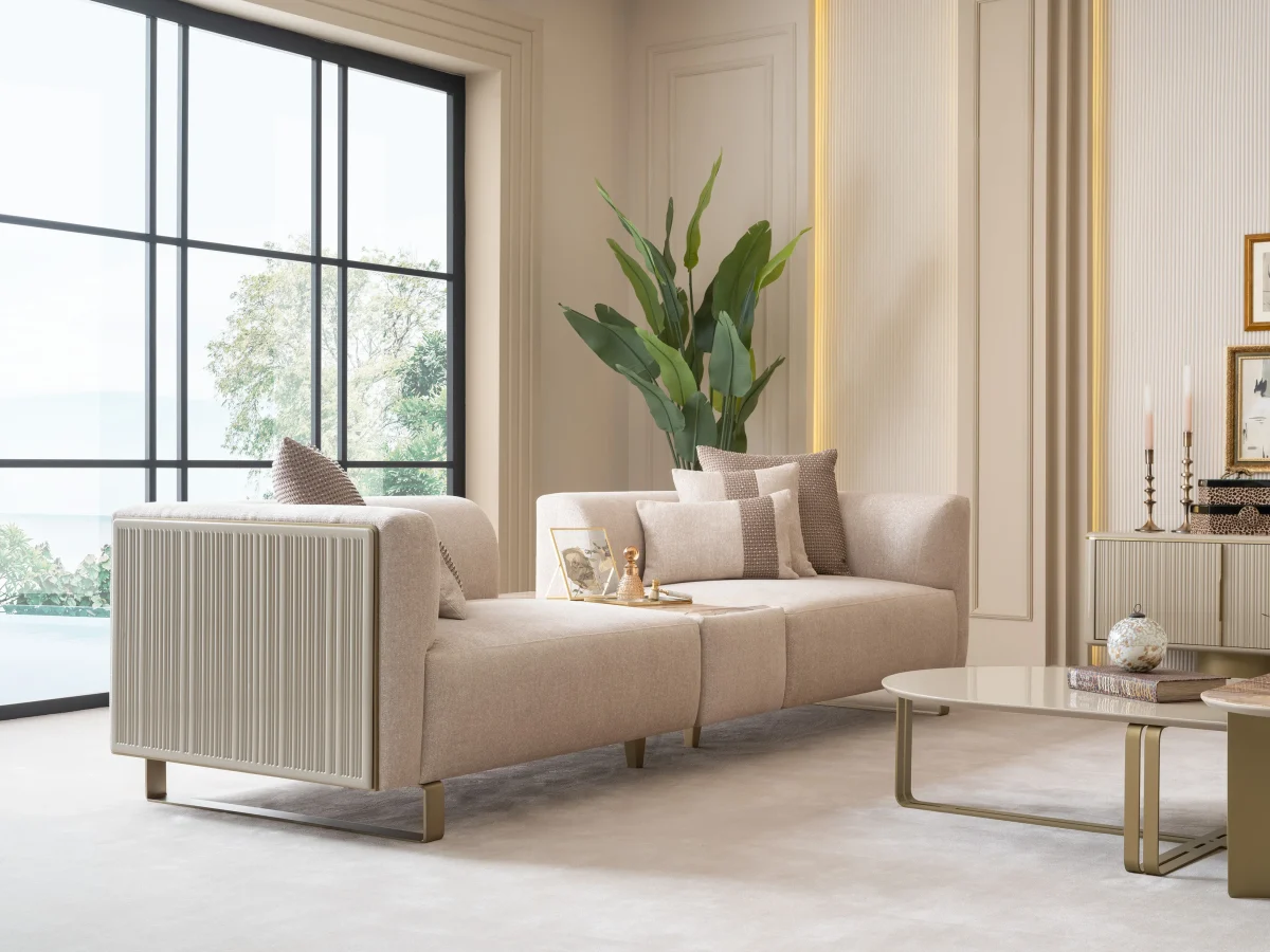 A luxurious modular Grandeur sofa set in a bright, modern living room. The set features cream-colored fabric upholstery with rectangular cushions and sleek gold metallic legs. Beside the modular sections sits a matching rounded armchair with gold accents. The scene includes minimalist decor, a marble coffee table, and large windows, highlighting the elegant design of the Grandeur furniture from Turkey.