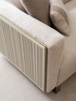 A luxurious modular Grandeur sofa set in a bright, modern living room. The set features cream-colored fabric upholstery with rectangular cushions and sleek gold metallic legs. Beside the modular sections sits a matching rounded armchair with gold accents. The scene includes minimalist decor, a marble coffee table, and large windows, highlighting the elegant design of the Grandeur furniture from Turkey.