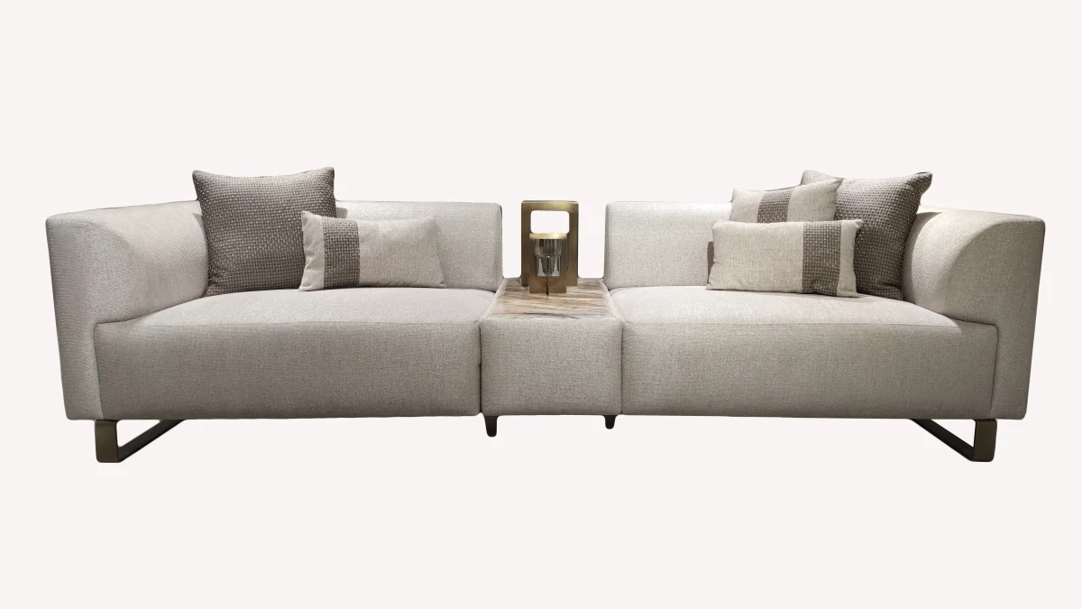 A luxurious modular Grandeur sofa set in a bright, modern living room. The set features cream-colored fabric upholstery with rectangular cushions and sleek gold metallic legs. Beside the modular sections sits a matching rounded armchair with gold accents. The scene includes minimalist decor, a marble coffee table, and large windows, highlighting the elegant design of the Grandeur furniture from Turkey.