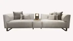 A luxurious modular Grandeur sofa set in a bright, modern living room. The set features cream-colored fabric upholstery with rectangular cushions and sleek gold metallic legs. Beside the modular sections sits a matching rounded armchair with gold accents. The scene includes minimalist decor, a marble coffee table, and large windows, highlighting the elegant design of the Grandeur furniture from Turkey.