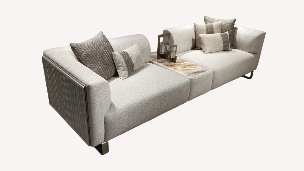 A luxurious modular Grandeur sofa set in a bright, modern living room. The set features cream-colored fabric upholstery with rectangular cushions and sleek gold metallic legs. Beside the modular sections sits a matching rounded armchair with gold accents. The scene includes minimalist decor, a marble coffee table, and large windows, highlighting the elegant design of the Grandeur furniture from Turkey.