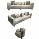 A luxurious modular Grandeur sofa set in a bright, modern living room. The set features cream-colored fabric upholstery with rectangular cushions and sleek gold metallic legs. Beside the modular sections sits a matching rounded armchair with gold accents. The scene includes minimalist decor, a marble coffee table, and large windows, highlighting the elegant design of the Grandeur furniture from Turkey.