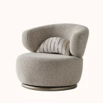 A modern Horizon Sofa set arranged in a chic living room. The furniture features textured grey upholstery with distinctive folded armrests and sleek champagne gold metallic legs. A matching round swivel armchair and a white coffee table with a cylindrical pouf complete the contemporary Turkish furniture ensemble.