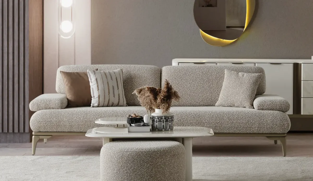 A modern Horizon Sofa set arranged in a chic living room. The furniture features textured grey upholstery with distinctive folded armrests and sleek champagne gold metallic legs. A matching round swivel armchair and a white coffee table with a cylindrical pouf complete the contemporary Turkish furniture ensemble.