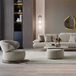 A modern Horizon Sofa set arranged in a chic living room. The furniture features textured grey upholstery with distinctive folded armrests and sleek champagne gold metallic legs. A matching round swivel armchair and a white coffee table with a cylindrical pouf complete the contemporary Turkish furniture ensemble.