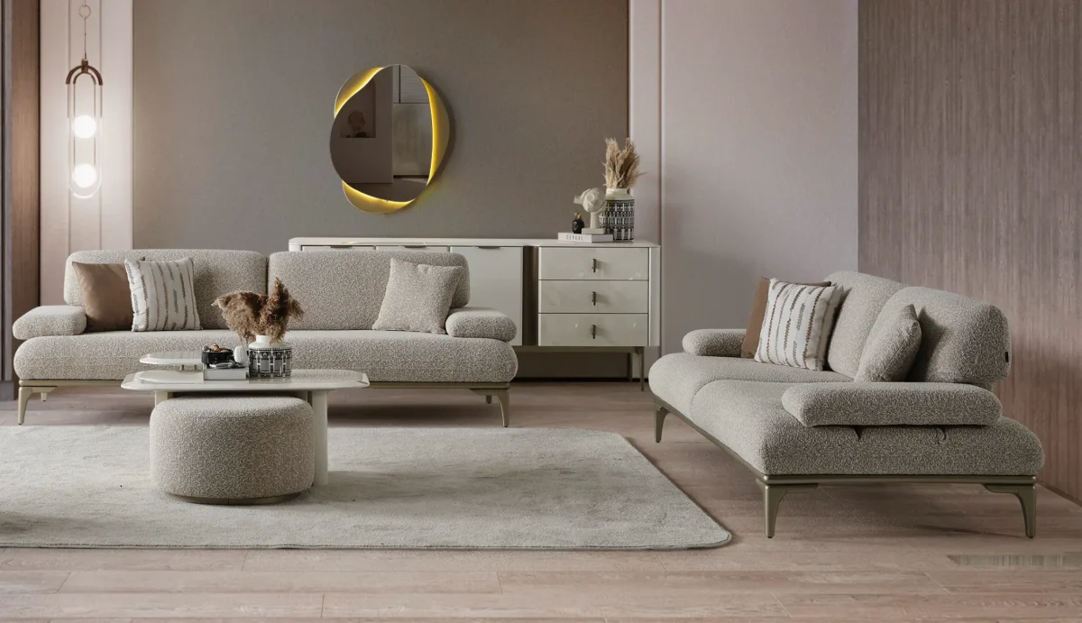 A modern Horizon Sofa set arranged in a chic living room. The furniture features textured grey upholstery with distinctive folded armrests and sleek champagne gold metallic legs. A matching round swivel armchair and a white coffee table with a cylindrical pouf complete the contemporary Turkish furniture ensemble.