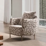 A modern accent armchair featuring an abstract geometric pattern upholstery, rounded continuous armrests, a matching throw cushion, and slim metallic tapered legs set in a brightly lit interior.
