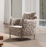A modern accent armchair featuring an abstract geometric pattern upholstery, rounded continuous armrests, a matching throw cushion, and slim metallic tapered legs set in a brightly lit interior.