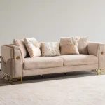 Luxia Sofa Set featuring a charcoal velvet three-seater, a blush pink loveseat with geometric patterns, and matching armchairs. The set is detailed with gold-toned metal bases and frames, set in a neoclassical room with arched windows and ornate wall moldings.