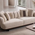 Sola Two Seater Sofa featuring rounded armrests, plush back cushions, and a wooden base trim in a modern living room setting.