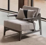 A contemporary Nova Armchair featuring an architectural open wooden frame, slim contoured armrests, a deep plush seat cushion, and a matching geometric accent pillow in a sunlit room setting.