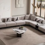 A modern modular Nova Corner Sofa featuring generous deep seating, multiple plush backrest cushions, geometric accent pillows, an ottoman extension, and low-profile metal legs against a plain white background.