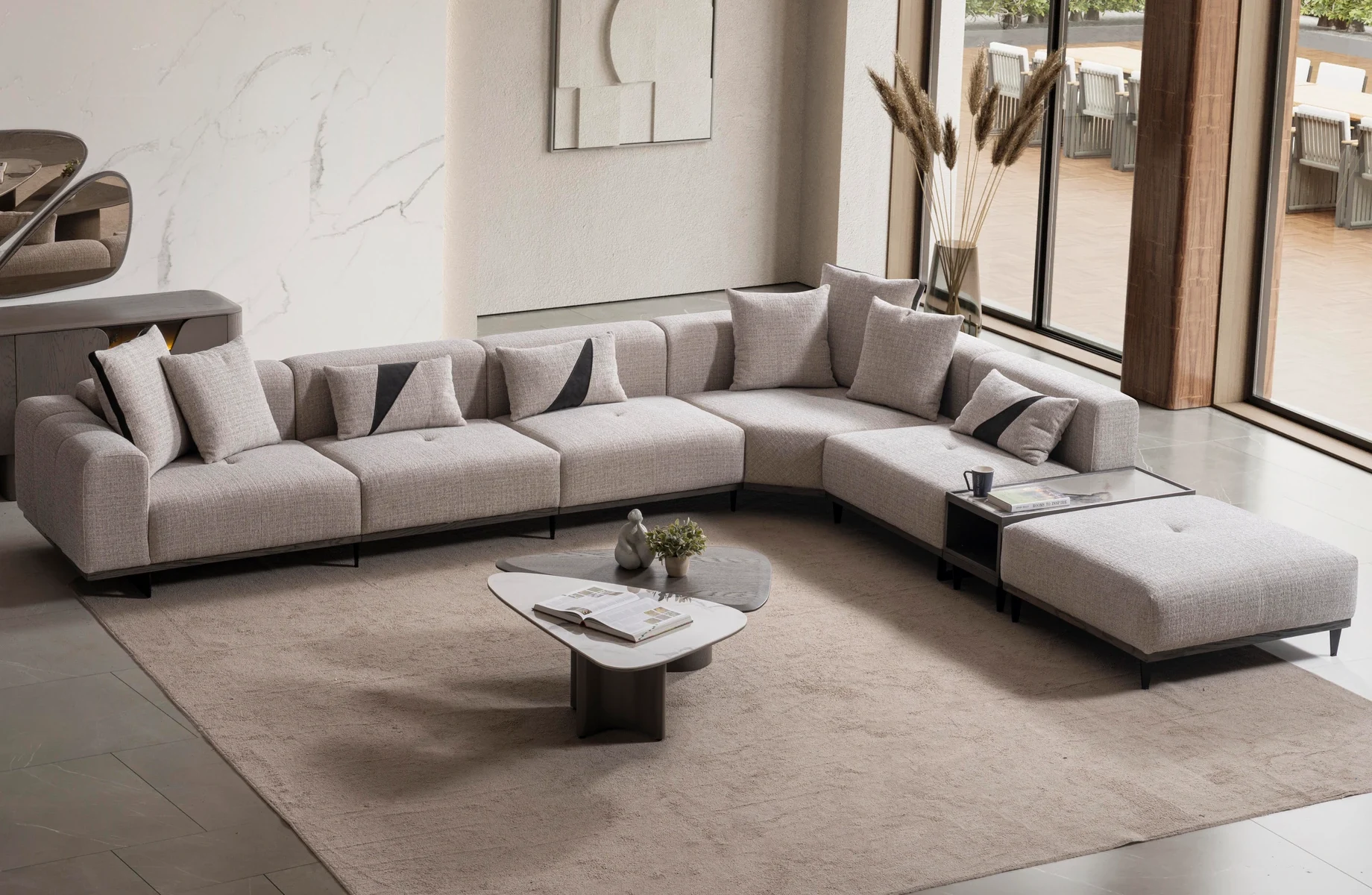 nova-corner-sofa-03 A modern modular Nova Corner Sofa featuring generous deep seating, multiple plush backrest cushions, geometric accent pillows, an ottoman extension, and low-profile metal legs against a plain white background.